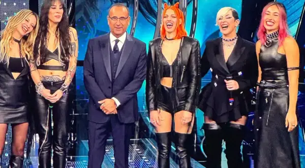 Micky Paiano con un completo scuro, affiancato da quattro donne con outfit in pelle audaci e colorati, sorridono sul palco.