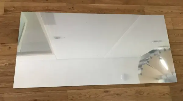 Un grand panneau de dibond miroir reflète le plafond et un mur blanc, posé sur un parquet en bois.