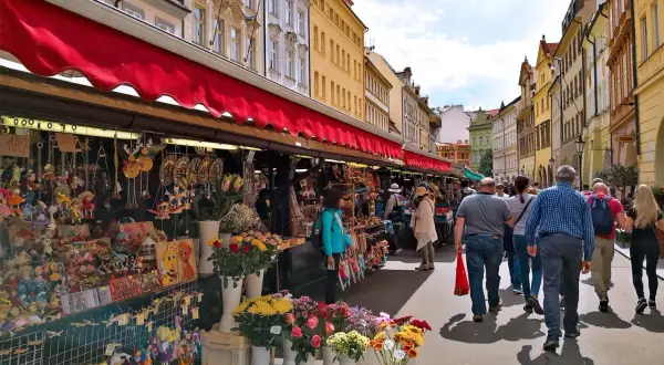 En livlig marknad i Prag med stånd fyllda av leksaker och blommor. Folk promenerar längs den soliga gatan.
