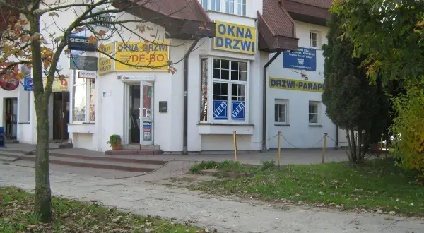 Budynek z żółtymi banerami "OKNA DRZWI" i "DRZWI PARAPETY", z drzewem po lewej.