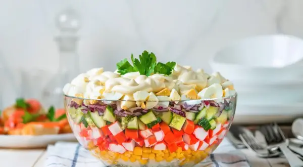Deliciosa ensalada de surimi en capas, con maíz, pepino, cebolla morada y huevo duro, cubierta de mayonesa y perejil.