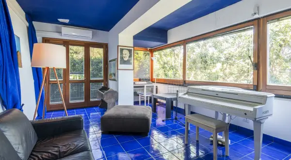 Interno con pavimento blu brillante, pianoforte bianco, divano nero e un ritratto di Padre Pio, come in testi lucio dalla la casa in riva al mare.