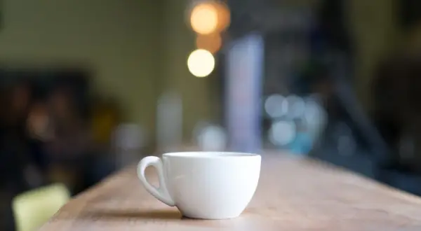 En vit, tom mått kaffekopp står på ett träbord. Bakgrunden är suddig med ljus.