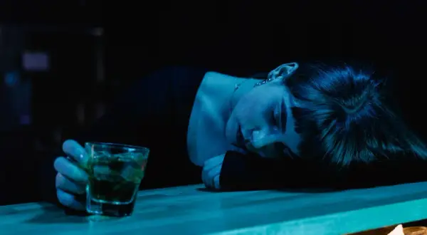 Zmęczona kobieta opiera głowę na blacie, obok szklanka z drinkiem. Mąż alkoholik obwinia mnie o wszystko, czuję się wyczerpana.