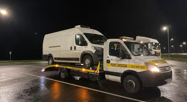 Nocne holowanie na drodze ekspresowej. Biała furgonetka jest transportowana na lawecie pomocy drogowej.
