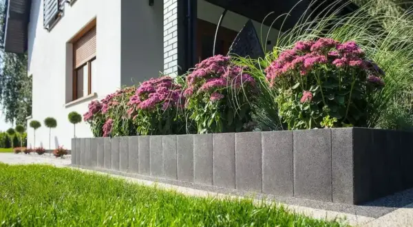 Nowoczesne donice z palisady przed domem, pełne kwitnących, różowych kwiatów i traw ozdobnych, tworzą piękny kontrast z bielą elewacji.