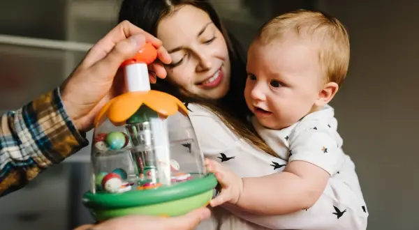 Mama i tata pokazują dziecku zabawkę. Maluch, choć nie jest jeszcze dwulatkiem, z zaciekawieniem kręci głową, obserwując kolorowe kulki.