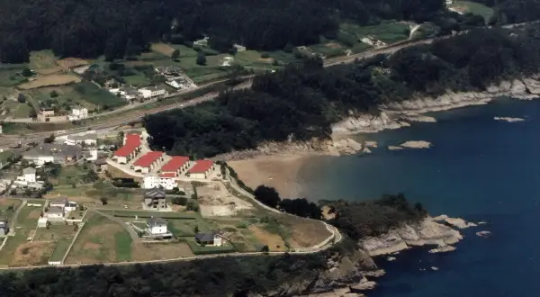 Vista aérea de casas con tejados rojos cerca de la playa de Sacido, rodeada de vegetación y el mar azul.