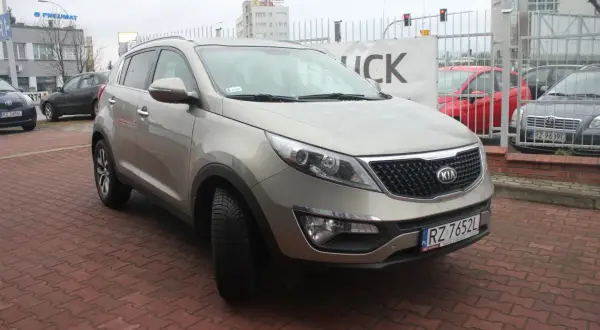 Złoty Kia Sportage 2012 na czerwonej kostce brukowej, z tablicą rejestracyjną RZ 7652L.