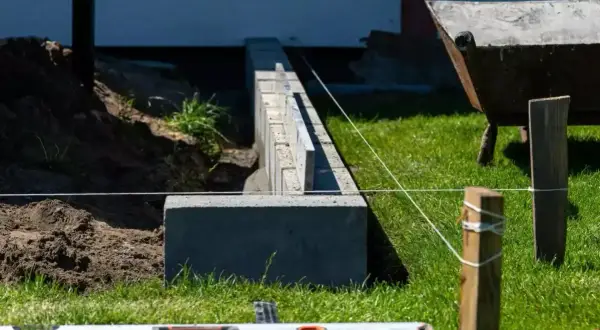 Budowa fundamentu pod ogrodzenie. Betonowe bloczki i poziomica wskazują precyzję prac. W tle taczka i kopiec ziemi.