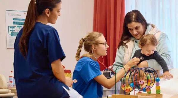 Ärzte in Krebszentren Deutschland untersuchen ein Kleinkind. Eine Krankenschwester hört mit dem Stethoskop ab, während eine Kollegin zusieht.