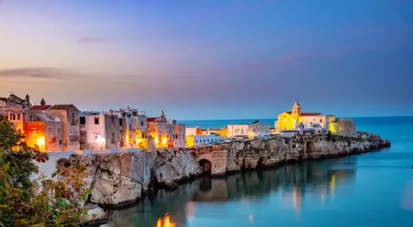 Vieste centro storico di sera, con case bianche illuminate e il mare blu che riflette le luci.