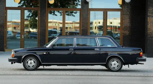 En mörkblå Volvo 264 TE, en lyxig limousin, står parkerad utanför en byggnad med stora fönster.