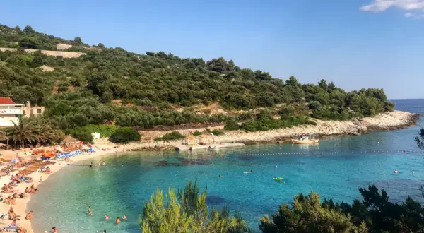 Słoneczna plaża na wyspie Vis, z turkusową wodą, zielonymi wzgórzami i ludźmi odpoczywającymi na piasku.