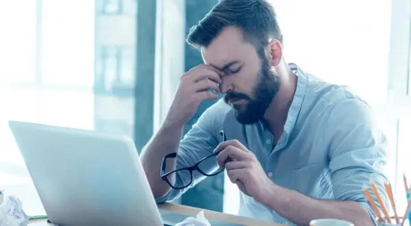 Mężczyzna z zarostem siedzi przy biurku, masując skronie. W ręku trzyma okulary, a przed nim leży laptop. Widać jego złe samopoczucie.