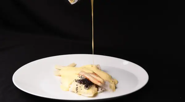 Ein Koch übergießt Fisch mit Spargel und Kaviar mit einer cremigen Sauce. Die Sauce sieht aus, als wäre die Sahne geronnen, was dem Gericht eine interessante Textur verleiht.