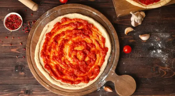 Une pâte à pizza crue est étalée sur une planche en bois, recouverte d'une sauce tomate maison. La question quelle sauce tomate pour pizza est au cœur de cette préparation.