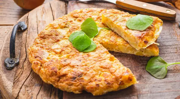 Goldbraune Tortilla de Patatas, garniert mit frischen Blättern, auf rustikalem Holzbrett. Dazu Zwiebeln und Besteck.