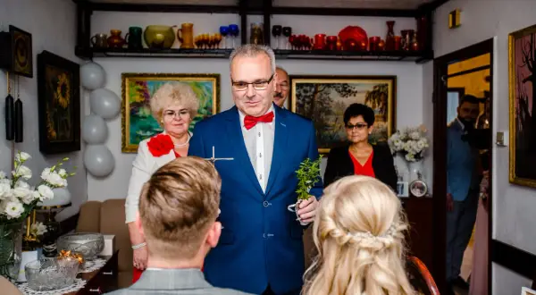 Mężczyzna w niebieskiej marynarce z czerwoną muszką dokonuje tradycyjne błogosławieństwo, trzymając krzyż i gałązkę.