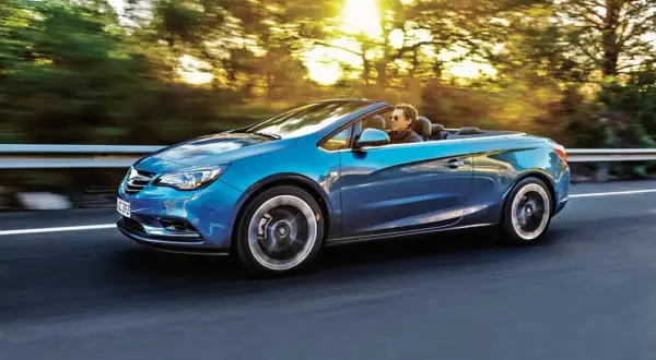 Niebieski Opel Cascada z otwartym dachem sunie po drodze, a słońce przebija się przez drzewa.