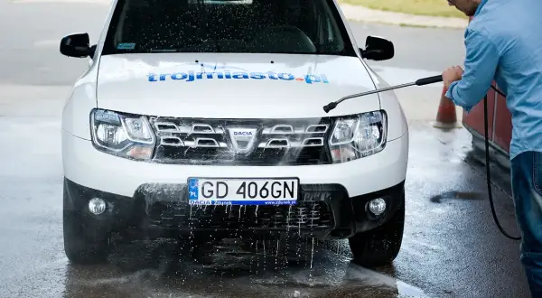 Mężczyzna myje białe Dacia Duster na myjni bezdotykowej, spryskując przód auta wodą pod ciśnieniem.