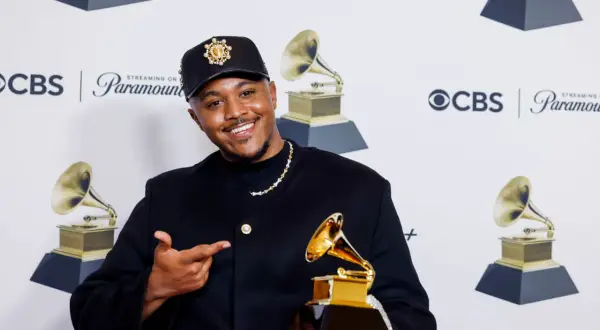 Khris Riddick-Tynes smiles, holding a Grammy award and pointing to it. He wears a black cap and jacket.