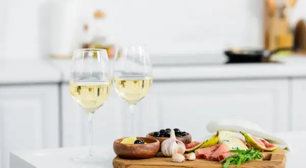 Due calici di vino bianco, ideali come vino bianco per cucinare, accompagnano un tagliere con formaggio, fichi, salumi e olive.