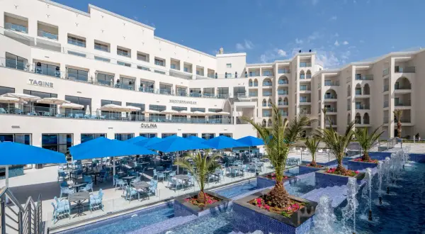 Luksusowy hotel z basenem i restauracją na świeżym powietrzu. Widać palmy, niebieskie parasole i stoły z krzesłami. Idealne miejsce na wakacje, jak na zdjęciach z **pickalbatros**.