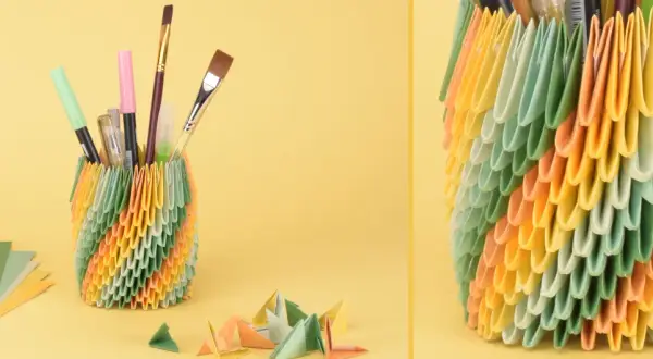 Bunter Papierkorb gefüllt mit Stiften und Pinseln, daneben Papierbögen und kleine Origami-Figuren.