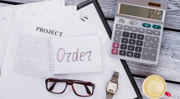 Na biurku leżą dokumenty, notatnik z napisem "Order", kalkulator, okulary, zegarek i kawa. To wszystko, co potrzebne do pracy, gdy zastanawiasz się, moq co to.