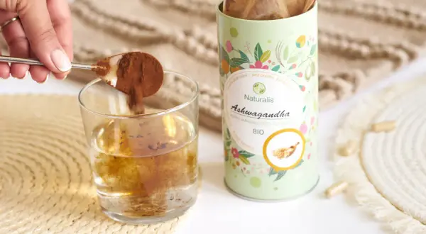 Łyżeczka z proszkiem ashwagandhy wsypywana do szklanki wody. Opakowanie Naturalis Ashwagandha BIO obok.