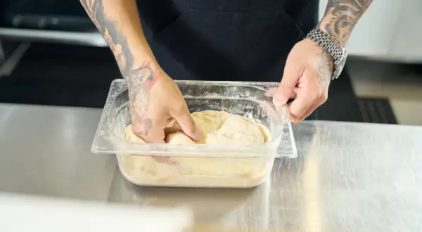 Ręce z tatuażami mieszają ciasto na pizzę w pojemniku. Poznaj prosty sposób, jak rozmrozić ciasto na pizzę, by było idealne.