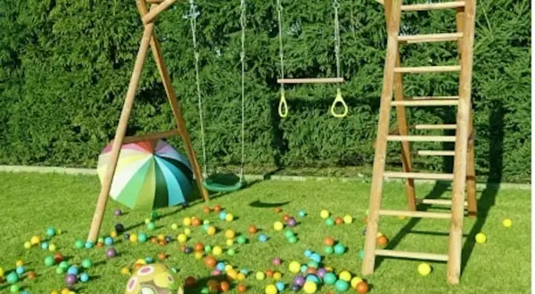 Drewniany plac zabaw ze zjeżdżalnią, huśtawką i drabinką, otoczony kolorowymi piłeczkami i parasolem.