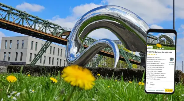 Moderne Skulptur vor Schwebebahn und Gebäude in Wuppertal. Ein Handy zeigt eine Tour durch Barmen, ein tolles Ziel für Ausflugsziele Wuppertal.