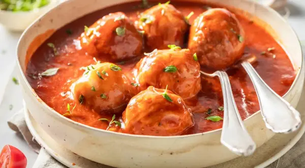 Albóndigas caseras con tomate frito, servidas en un cuenco de cerámica con hierbas frescas y dos cucharas listas para disfrutar.