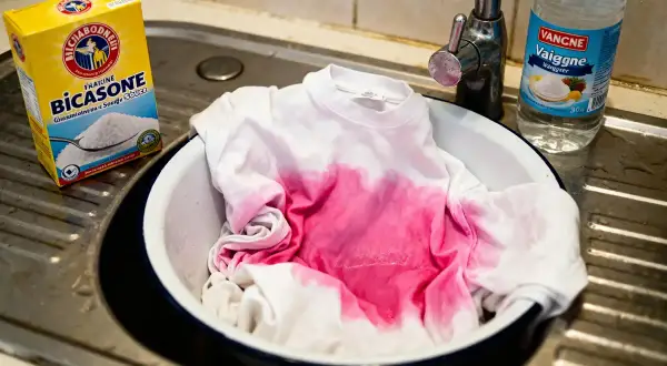 Un t-shirt blanc taché de rose trempe dans une bassine. À côté, une boîte de bicarbonate de soude et une bouteille de vinaigre, une astuce grand-mère pour le linge déteint.