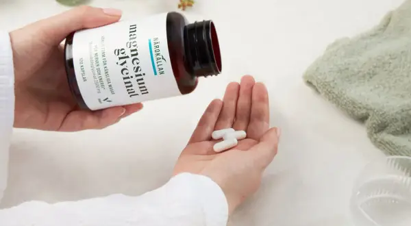 Händer häller upp tre kapslar magnesiumglycinat ur en flaska. Ett uppiggande medicin för nerver och energi.
