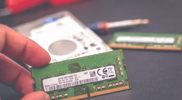 Dłoń trzyma moduł pamięci RAM 8 GB. Obok leży dysk twardy i drugi moduł RAM, sugerując wybór: 8 czy 16 GB RAM w laptopie.