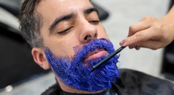 Un homme se fait teindre la barbe en bleu vif par un professionnel. La couleur est appliquée avec une brosse.