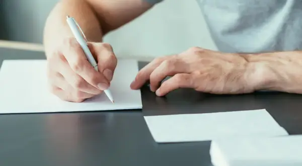 Dłoń pisząca długopisem na białej kartce papieru, obok koperty.