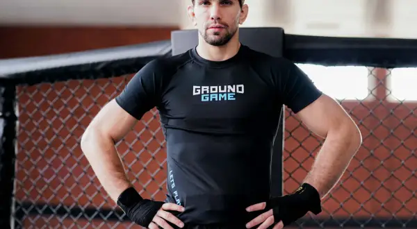 Mężczyzna w czarnym rashguardzie z napisem "GROUND GAME" stoi w klatce. Czy to jest to, co nazywamy rashguard co to?