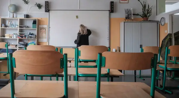 Klasa szkolna z pustymi ławkami. Nauczycielka pisze na tablicy. Zastanawiasz się, ile zarabia pomoc nauczyciela na pół etatu?