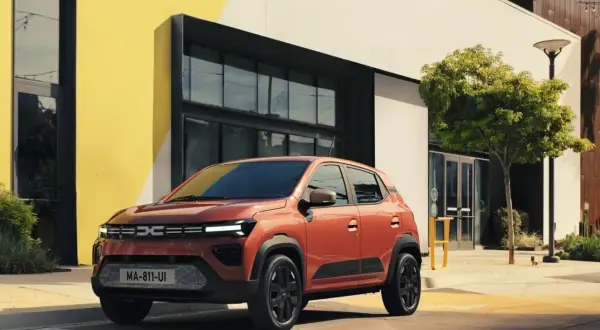 Pomarańczowy Dacia Spring Electric zaparkowany przed nowoczesnym budynkiem z żółtymi akcentami.
