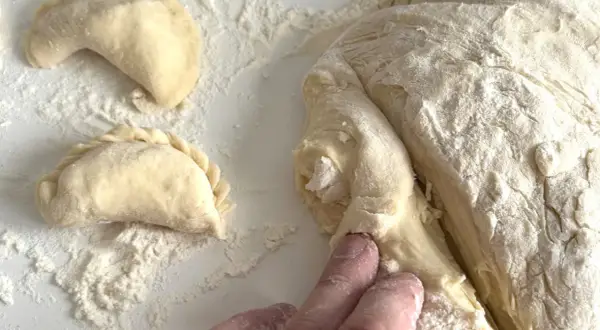 Ręce formują ciasto na pierogi, obok leżą już ulepione pierożki.