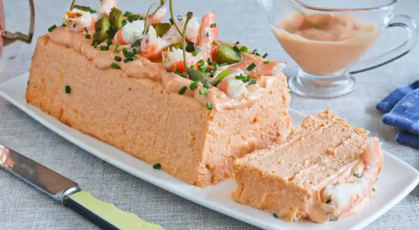 Pastel de pescado decorado con gambas, pepinillos y alcaparras. Una porción ya cortada revela su interior.