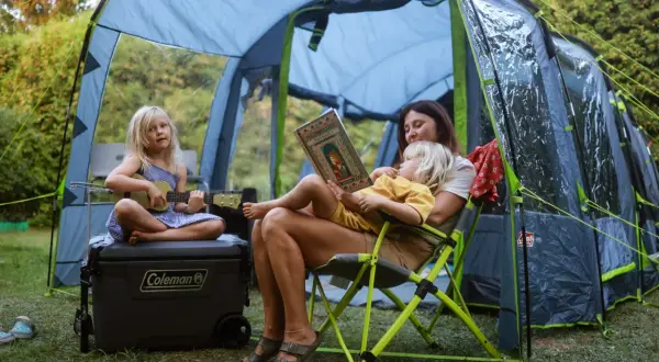Rodzina odpoczywa pod namiotem podczas festiwalu słońca. Dziecko gra na ukulele, a mama czyta książkę.