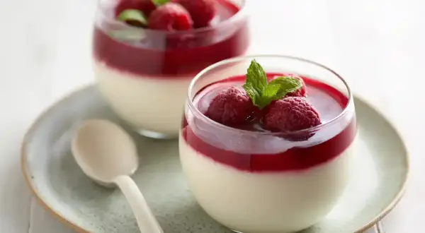 Två glas med krämig pannacotta med hallonspegel, toppad med färska hallon och mynta. En ljus sked ligger på fatet.