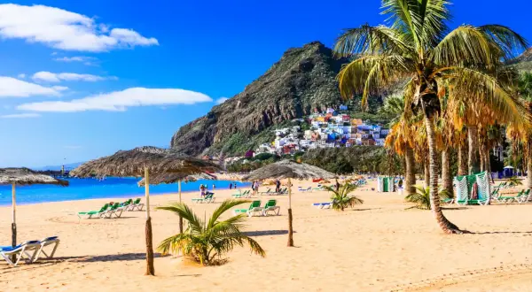 Plage de sable doré, palmiers, parasols et maisons colorées perchées sur une colline. Une invitation à visiter Tenerife pour des vacances de rêve.
