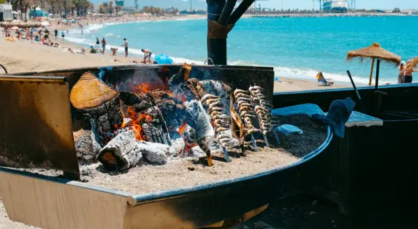 Pescado a la brasa en un chiringuito del paseo marítimo de Málaga. El mar y la playa con gente al fondo.