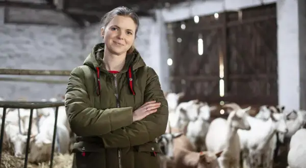 Kobieta w zielonej kurtce stoi na tle stada kóz. Jej uśmiech sugeruje, że hodowla kóz opłacalność jest dla niej satysfakcjonująca.